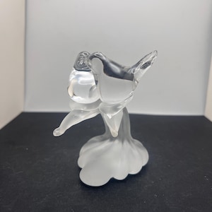 Può includere: Una scultura in vetro trasparente di un uccello posato su un fiore. L'uccello ha un corpo arrotondato e ali spiegate. Il fiore ha una base a forma di petalo. La scultura è realizzata in vetro trasparente con dettagli smerigliati.