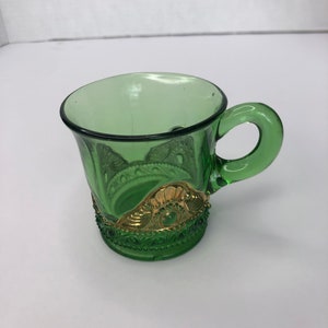 Puede incluir: Una taza de vidrio verde con un borde dorado y un asa. La taza tiene un diseño decorativo en el lateral.