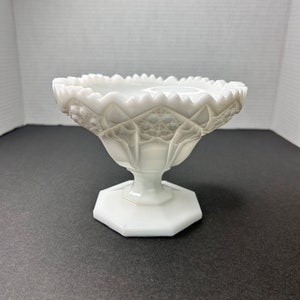 Puede incluir: Un cuenco blanco de vidrio de leche sobre un pedestal con un borde festoneado y un diseño de corte complejo. El cuenco tiene una base hexagonal.
