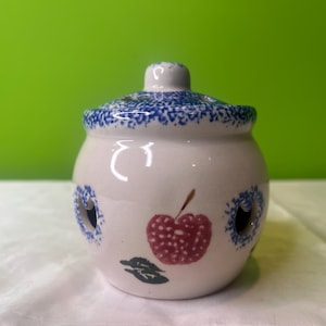 Op de afbeelding: Witte keramische pot met een blauw-wit gespikkelde deksel. De pot heeft een blauw-witte rand en een rood appelontwerp. De pot heeft gaten aan de zijkanten.