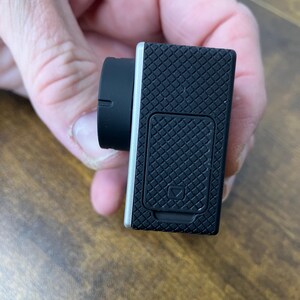 May include: Black action camera with a silver trim and a black lens. The camera has a textured, grid-like pattern on the back and a small door with a square symbol. The lens is round and black.