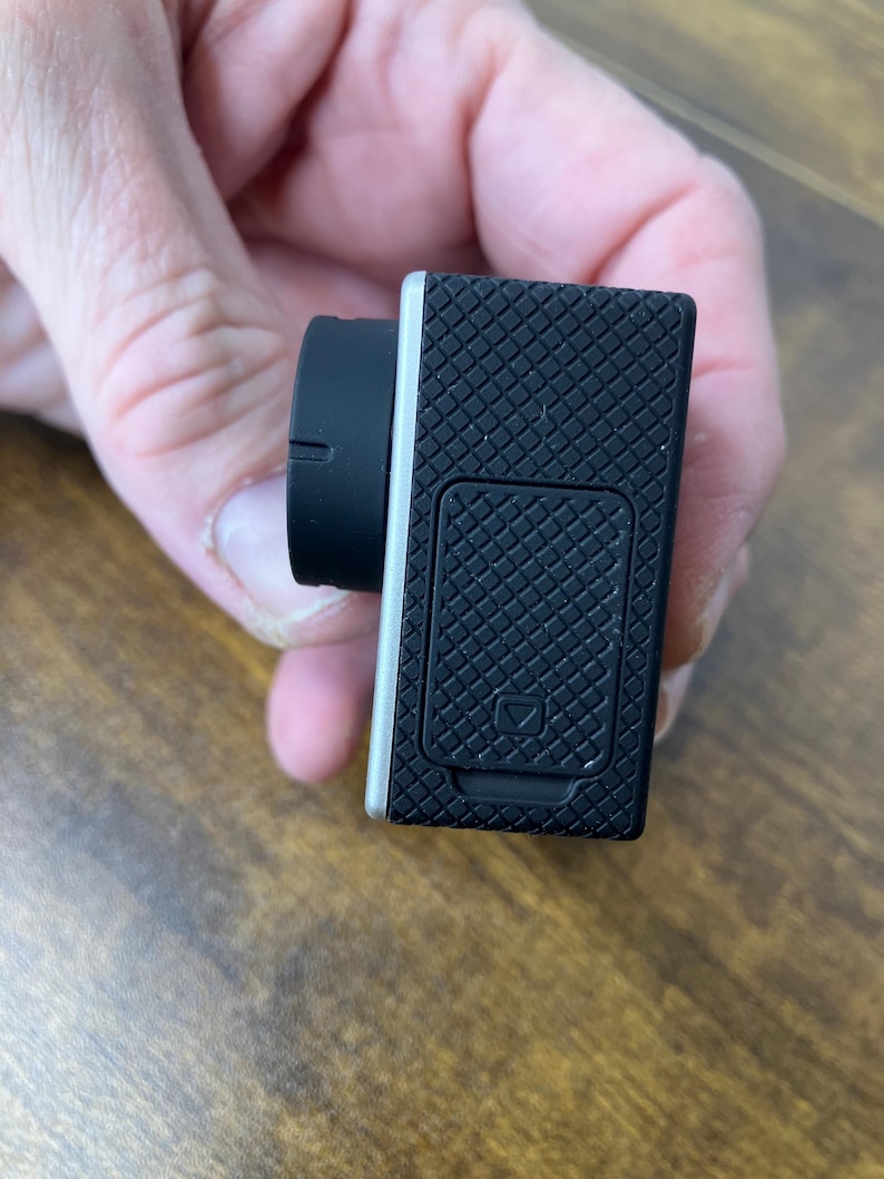 May include: Black action camera with a silver trim and a black lens. The camera has a textured, grid-like pattern on the back and a small door with a square symbol. The lens is round and black.