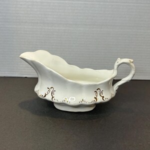 May include: White ceramic gravy boat with a scalloped rim and a decorative gold pattern. The gravy boat has a handle and a small footed base. A classic serving dish for sauces and gravies.