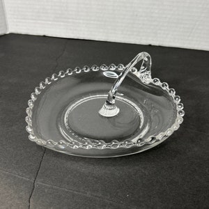 Puede incluir: Un plato de servir de vidrio transparente con un borde de cuentas y un asa. El plato es redondo y tiene un pequeño borde alrededor del borde.