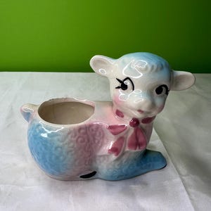 Puede incluir: Una maceta de cordero de cerámica en azul pastel y rosa. El cordero tiene una cara blanca con ojos negros y un lazo rosa. La maceta tiene una abertura redonda para plantas o objetos pequeños. Una pieza decorativa encantadora.