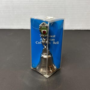 May include: A silver bell with a green and red apple design on the top. The bell is in a clear plastic box with a blue background. The box says "The Great American Collectors Bell".