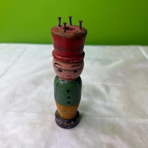 Könnte beinhalten: Vintage Strickliesel-Puppe aus Holz mit rotem Hut, grüner Jacke und gelber Hose. Die Puppe hat ein bemaltes Gesicht und sechs Metallnägel auf der Oberseite. Der Sockel ist schwarz und die Puppe steht auf einer weißen Oberfläche.