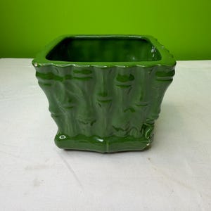 Puede incluir: Una maceta cuadrada de cerámica verde oscuro brillante. La maceta tiene un diseño texturizado similar al bambú en los lados y una abertura cuadrada en la parte superior. La maceta está sobre una superficie blanca contra un fondo verde.