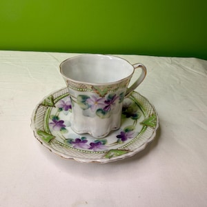 May include: A white teacup and saucer set with a floral design. The cup and saucer feature purple and green flowers with gold trim. The saucer has a scalloped edge. The teacup is sitting on the saucer.