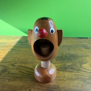 May include: Wooden nutcracker in the shape of a clown's head. The clown has a red nose, white eyes, and small ears. The mouth is open, revealing the nut-cracking mechanism. The nutcracker is brown and stands on a wooden base.