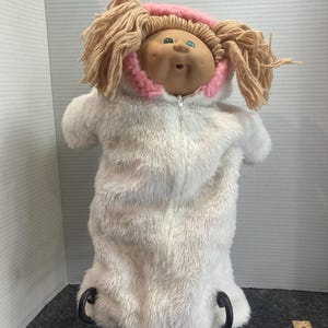 Könnte beinhalten: Eine Puppe mit beigem Vinylgesicht, grünen Augen und hellbraunem Wollhaar, bekleidet mit einem weißen Kunstfell-Kapuzenanzug mit rosa Innenfutter und Reißverschluss. Die Puppe steht auf einem schwarzen Metallständer neben einem Holzlineal.