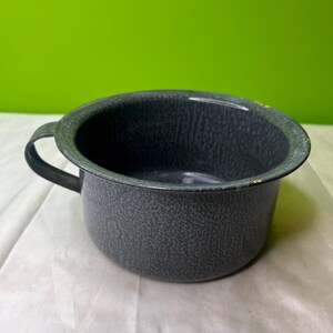 May include: A speckled, dark gray enamelware chamber pot with a single handle. The pot has a rounded shape and a slightly flared rim. The interior is a solid dark gray. The background is a solid green.