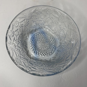 May include: A clear glass bowl with a floral pattern. The bowl has a circular shape with a detailed embossed design of flowers and leaves around the rim. The centre of the bowl features a textured starburst pattern.