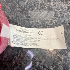 May include: A white fabric product tag attached to a pink plush item. The tag features black text including The Walt Disney Company logo, CE mark, and manufacturing details such as Made in China, polyester fiber, and plastic pellets content.
