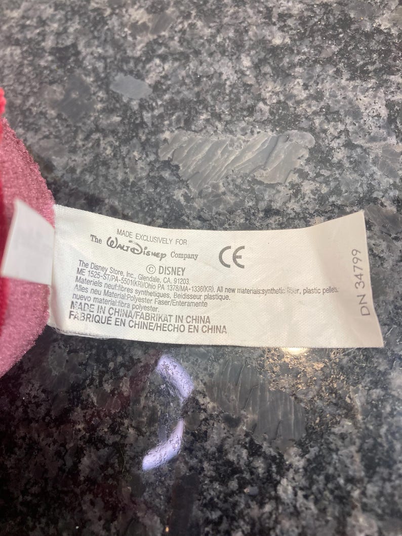 May include: A white fabric product tag attached to a pink plush item. The tag features black text including The Walt Disney Company logo, CE mark, and manufacturing details such as Made in China, polyester fiber, and plastic pellets content.