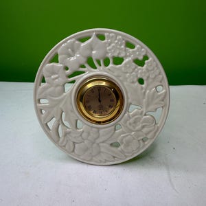 May include: A cream-colored decorative clock with a gold-tone clock face. The clock is round with a floral and leaf design cut out around the clock face. The clock face has Roman numerals.