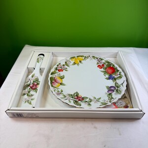 May include: A white ceramic cake plate and server set with a fruit and floral design. The plate has a scalloped edge and a gold rim. The server has a matching design. The set is in a white box.