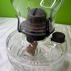 Puede incluir: L&aacute;mpara de aceite de vidrio transparente con tapa y detalles de metal oscuro. La l&aacute;mpara tiene una base redonda y un gran globo de vidrio transparente. La mecha es visible dentro del dep&oacute;sito de vidrio. La l&aacute;mpara est&aacute; dise&ntilde;ada para aceite.