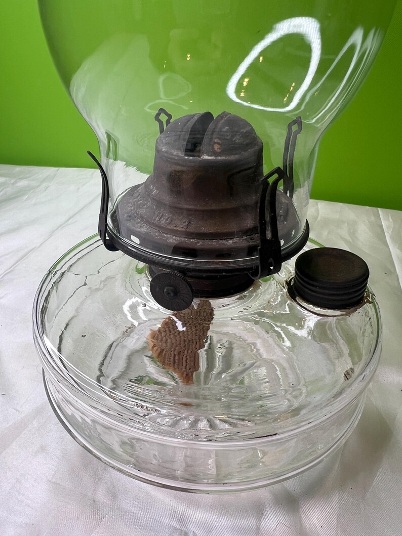 Puede incluir: L&aacute;mpara de aceite de vidrio transparente con tapa y detalles de metal oscuro. La l&aacute;mpara tiene una base redonda y un gran globo de vidrio transparente. La mecha es visible dentro del dep&oacute;sito de vidrio. La l&aacute;mpara est&aacute; dise&ntilde;ada para aceite.