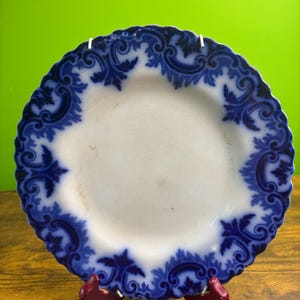 May include: A round ceramic plate with a scalloped edge featuring a white center and a wide, dark blue border decorated with intricate scrolling leaf patterns. The surface of the plate shows subtle signs of age with light discoloration.