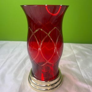 Puede incluir: Un jarrón de cristal rojo rubí con base dorada. El jarrón tiene una parte superior acampanada y un patrón de diamantes grabado en el cristal. La base tiene una serie de anillos concéntricos. El jarrón está sobre una superficie blanca.