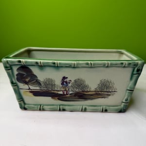 Puede incluir: Jardinera rectangular de cerámica con un borde verde claro estilo bambú. La jardinera presenta una escena de paisaje pintada con árboles y una figura. El interior es blanco y el exterior es de color verde claro.