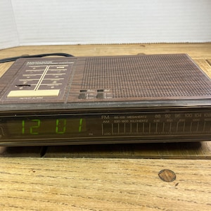 May include: A vintage Magnavox digital alarm clock with a brown wood-grain finish. The clock displays the time in green digits and has FM radio capabilities. The clock has a rectangular shape and a speaker grill.