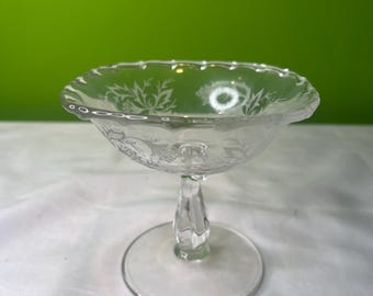 May include: Clear glass pedestal bowl with etched floral designs. The bowl has a scalloped rim and a round base. The stem is twisted. The bowl is likely used for serving desserts or displaying small items.