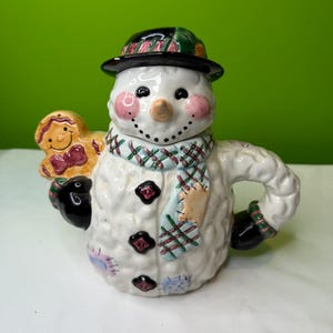 May include: A ceramic snowman figure wearing a black hat, a scarf, and holding a gingerbread man. The snowman has rosy cheeks, a carrot nose, and black button eyes. The scarf is green, blue, and red. The gingerbread man is holding a heart.