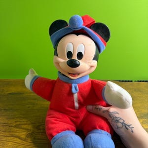 May include: A vintage Mickey Mouse plush doll wearing a red onesie with blue accents on the feet, cuffs, and hat. The doll has a zipper down the front and white gloves. The hat is red and blue with a blue ball on top. The doll is smiling.