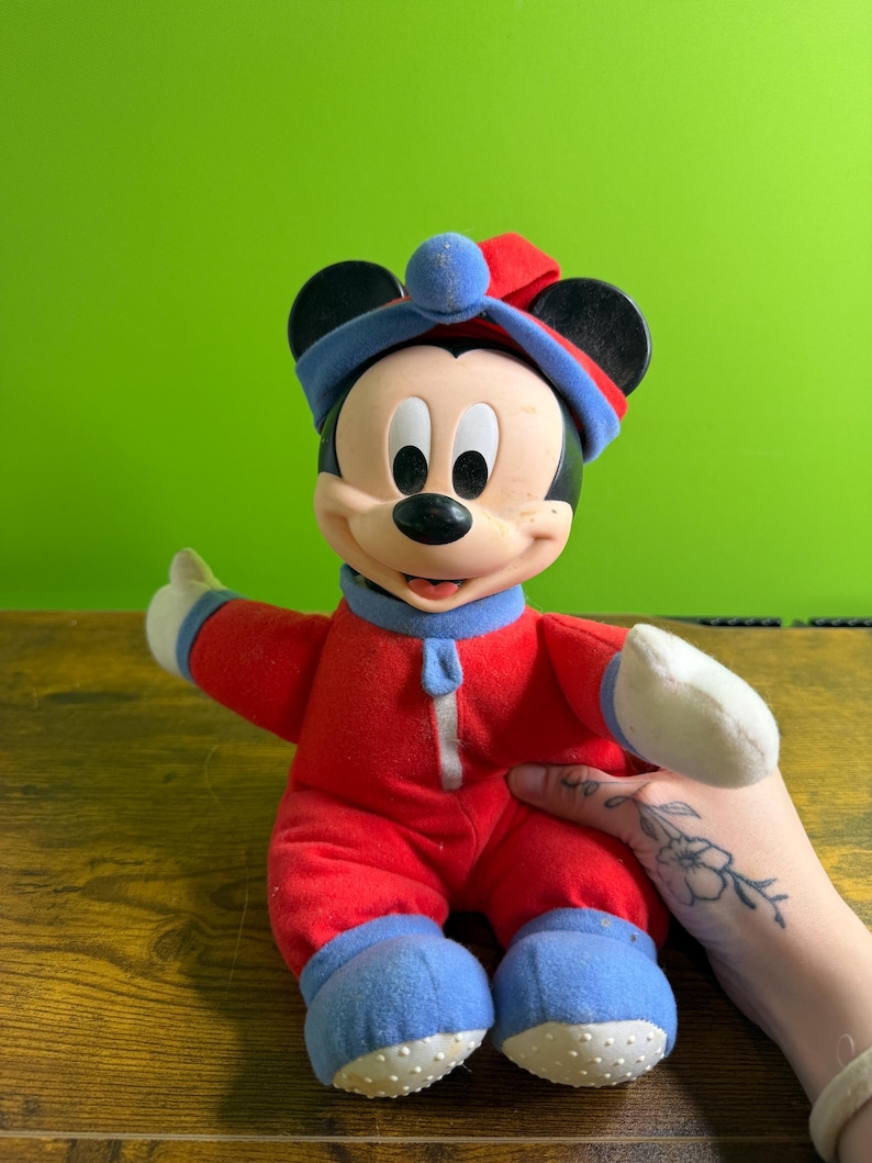 May include: A vintage Mickey Mouse plush doll wearing a red onesie with blue accents on the feet, cuffs, and hat. The doll has a zipper down the front and white gloves. The hat is red and blue with a blue ball on top. The doll is smiling.