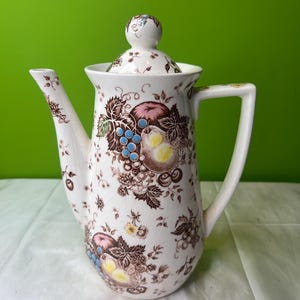 Puede incluir: Una cafetera de cerámica blanca con un patrón floral marrón y verde con frutas y uvas. La cafetera tiene una tapa con un pomo en la parte superior.