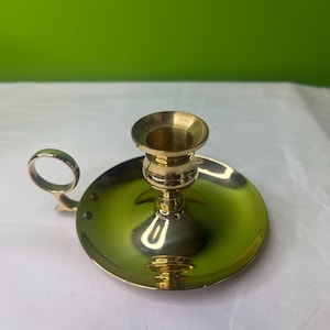 May include: A brass candlestick holder with a circular base and a small handle. The holder has a cup-shaped top to hold a candle. The brass has a shiny, reflective surface. The background is a solid green.