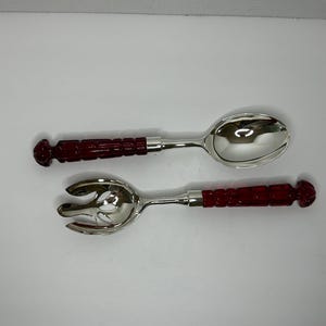 May include: A set of two silver salad serving utensils with red plastic handles. The spoon has a rounded bowl and the fork has a slotted design.