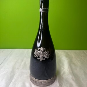 May include: A dark green glass bottle with a silver emblem and base. The bottle has a long neck and a rounded body. The silver emblem is ornate and features a crest design. The base of the bottle has engraved text.