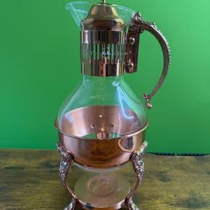 Op de afbeelding: Een vintage koffiezetapparaat van helder glas en koper, met een decoratieve handgreep en voet. Het koffiezetapparaat heeft een heldere glazen karaf en een koperkleurig metalen frame. De handgreep en voet hebben sierlijke details.