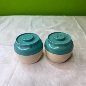 May include: A pair of vintage salt and pepper shakers. Each shaker is two-toned, with a cream-colored base and a teal-colored top. The tops have small holes for dispensing the contents.