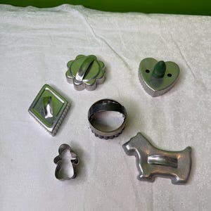 Könnte beinhalten: Eine Sammlung von Vintage-Keksformen aus Metall in verschiedenen Formen, darunter eine Raute, eine Blume, ein Herz, ein Kreis, ein Lebkuchenmann und ein Hund. Die Ausstecher sind silberfarben und auf einer weißen Oberfläche angeordnet.
