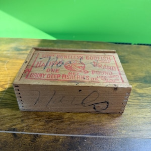 May include: A vintage wooden box with red text reading "Boneless Codfish Brand" and "Briny Deep Fisheries Ltd." The box also features the word "Halifax" and the word "Miles" written on the side.