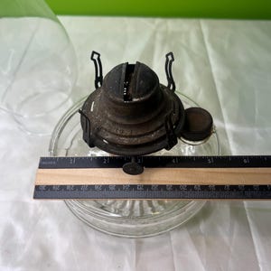 Puede incluir: Un quemador de l&aacute;mpara de aceite antiguo con una base de vidrio transparente. El quemador de metal es de color marr&oacute;n oscuro y tiene una peque&ntilde;a tapa redonda. Una regla muestra que la base tiene aproximadamente 12,7 cm de di&aacute;metro.
