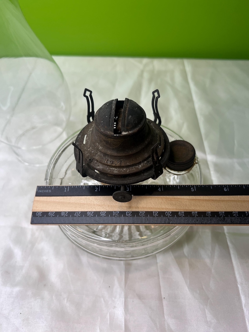 Puede incluir: Un quemador de l&aacute;mpara de aceite antiguo con una base de vidrio transparente. El quemador de metal es de color marr&oacute;n oscuro y tiene una peque&ntilde;a tapa redonda. Una regla muestra que la base tiene aproximadamente 12,7 cm de di&aacute;metro.