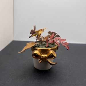 Può includere: Un piccolo vaso di ceramica bianca con un dettaglio a fiocco dorato, contenente fiori metallici in miniatura nei toni dell'oro, del rosso e del marrone. Il vaso ha un bordo dorato. Il tema generale è decorativo e ornamentale.