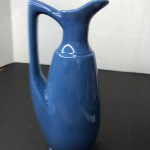 Puede incluir: Una jarra de cerámica azul con un asa curva única. La jarra tiene un acabado liso y brillante y una base redondeada.
