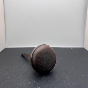 May include: A vintage, round, metal oil can with a long spout. The can is a dark, mottled brown colour, with signs of wear and age. The spout extends from the side of the can.