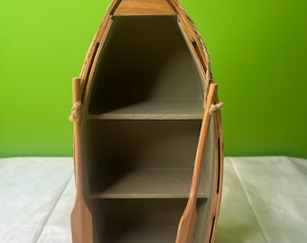 May include: A wooden boat-shaped shelf unit with three shelves inside. The unit has a natural wood finish, with rope detailing. Two wooden oars are attached to the sides. The shelf is designed to resemble a small boat.