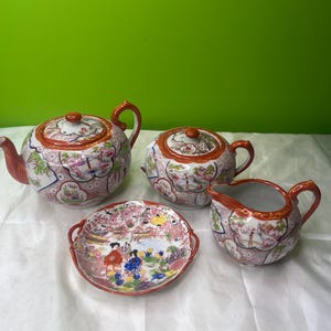 May include: A vintage porcelain tea set with a teapot, creamer, sugar bowl, and serving plate. The set features a floral and landscape design with red, pink, and blue accents. The teapot and sugar bowl have lids with red knobs.