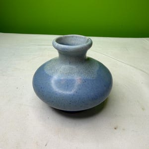 Puede incluir: Un pequeño jarrón redondo de cerámica con un esmalte moteado azul y gris. El jarrón tiene un cuerpo bulboso y un cuello corto y acampanado. El degradado de color pasa de gris claro en la parte superior a azul más oscuro en la base.