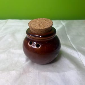 Può includere: Un piccolo barattolo rotondo in ceramica marrone con un tappo di sughero. Il barattolo ha due piccoli fori vicino alla parte superiore e una finitura lucida. Il tappo è di colore marrone chiaro e si adatta perfettamente sopra il barattolo. Il barattolo è probabilmente per conservare spezie o altri piccoli oggetti.
