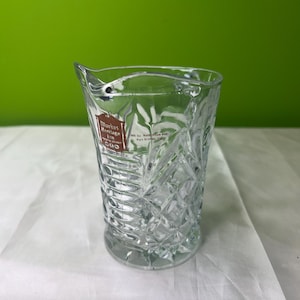 May include: Clear glass cocktail mixing glass with a diamond pattern and a pouring spout. A red and white label is visible on the front of the glass. The glass is sitting on a white surface with a green background.