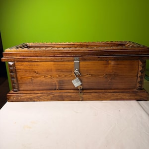 Puede incluir: Un cofre de madera con cerradura y llave. El cofre está teñido de un color marrón cálido y tiene adornos decorativos en la parte superior. La cerradura es plateada y la llave está en la cerradura. El cofre está sobre una superficie blanca.